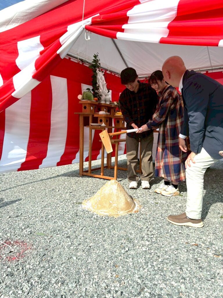 紅白幕が張られた晴天の地鎮祭会場で、施主が砂盛に対して木槌で鍬入れの儀を行う様子。祭壇にお供え物が並び、家づくりの工事の安全と繁栄を祈願する伝統的な儀式が執り行われています。