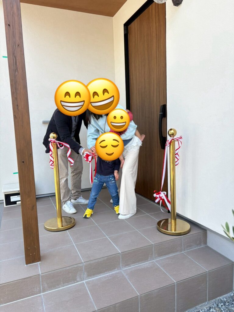 木目調の玄関ドアとタイル仕上げのポーチが映える新居の玄関前で、笑顔でテープカットを行うご家族。引き渡し式の輝かしい一瞬が、これからの安心で快適な暮らしを象徴している。