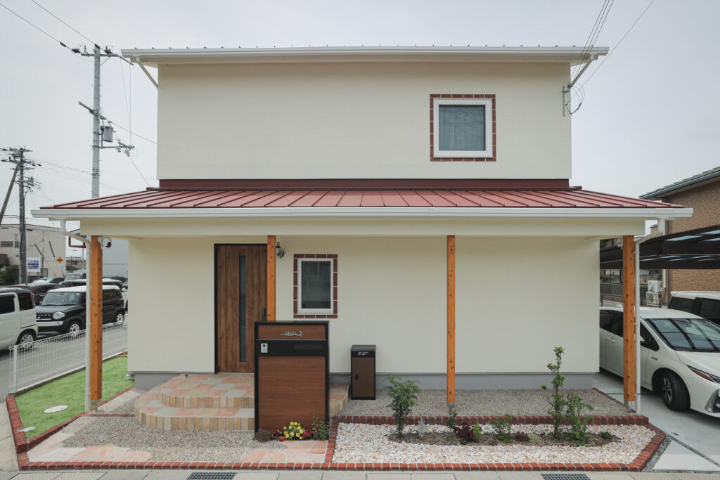 クリーム色の塗り壁に赤い金属屋根と木製の柱が温かみを添える、南欧風のナチュラルな外観。