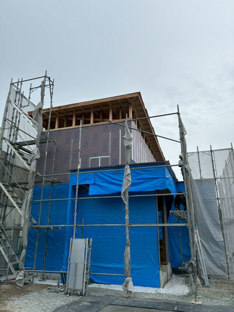 足場が組まれ、木造の構造体の上層階に透湿防水シートが施工され、下層階がブルーシートで保護されている建築中の住宅。
