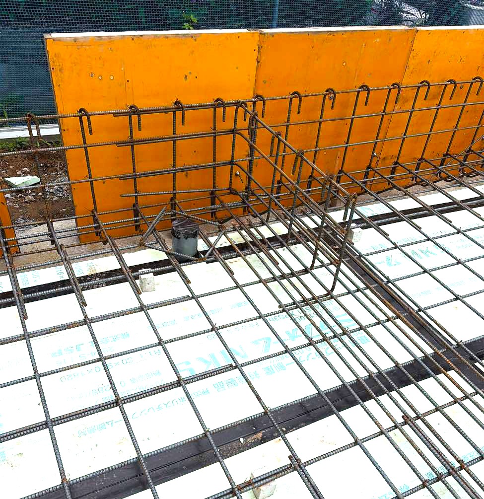 住宅の基礎工事現場にて、木製の型枠が組まれ、防湿シートと配筋が施されたベタ基礎の施工風景を、作業員がチェックしています。