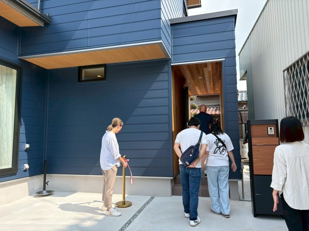 深みのあるネイビーのガルバリウム外壁に木目の軒天がアクセントとなった、モダンな住宅の玄関前。見学会のような雰囲気の中、訪問者が入り口へ向かう様子が伺える外観。