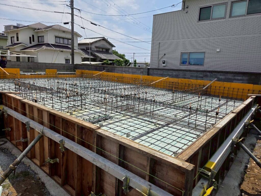 住宅の基礎工事現場にて、防湿シートと断熱材が敷かれた上に鉄筋が組まれ、施工の寸法精度を確認するためにスケールを当てて測定している様子。