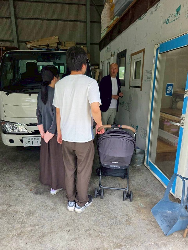 倉庫や作業場のような空間で、ベビーカーを押す夫婦が工務店の担当者から建材や設備の説明を受けている打ち合わせの様子。