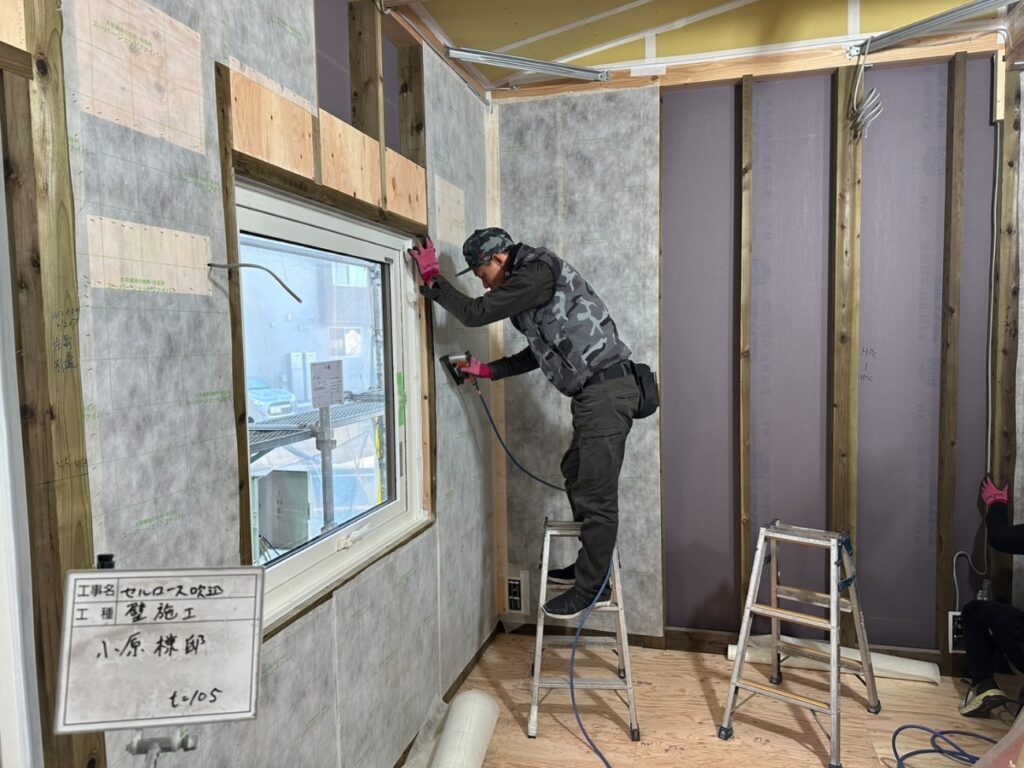 木造住宅の建築現場にて、職人が踏み台に乗り、気密・断熱層を施工するための壁下地材をエアタッカーで丁寧に固定している様子。