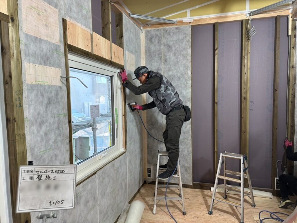 木造住宅の建築現場にて、職人が踏み台に乗り、セルロースファイバー断熱のための気密シートを窓枠周りにエアタッカーで丁寧に固定している様子。