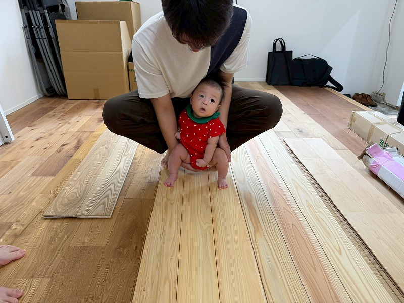 無垢材のフローリングが施工中の室内で、赤ちゃんを抱えたお父さんが床材の質感を確認している様子。