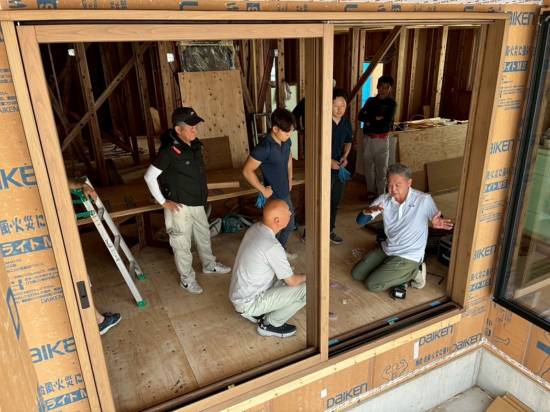 木造軸組と耐力面壁で施工された建築中の室内にて、大開口の窓枠の納まりを確認しながら打ち合わせを行うスタッフたち。