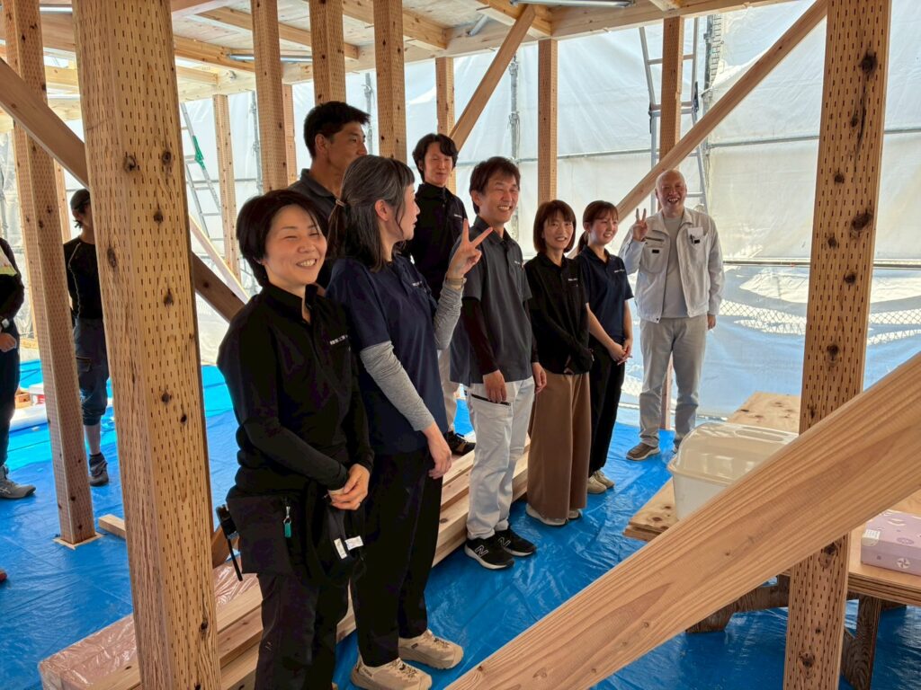 木造軸組構造の柱や梁が組み上がった建築中の現場にて、笑顔で並んで記念撮影に臨むスタッフたち。