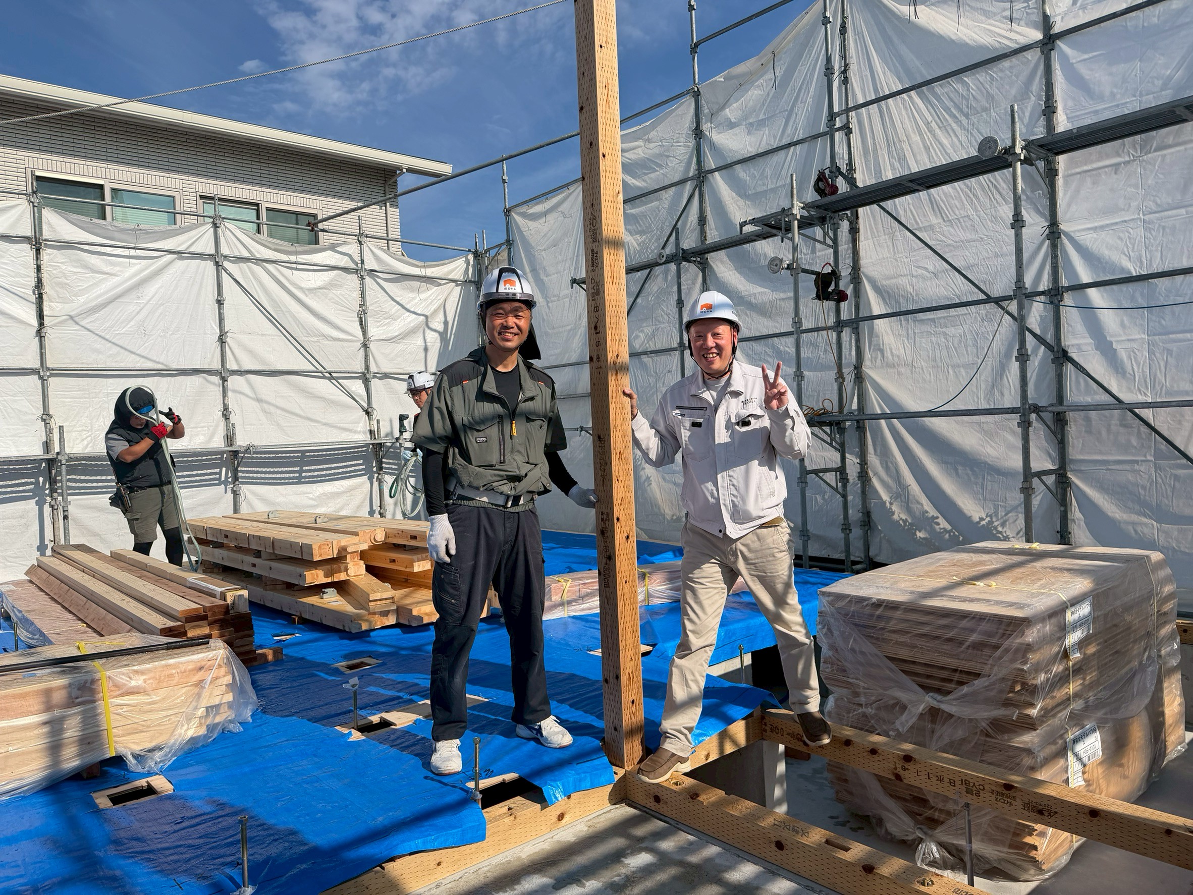 足場と養生シートに囲まれた建築中の現場で、木造軸組の柱や構造材の傍らに立ち笑顔を見せる作業員たち。
