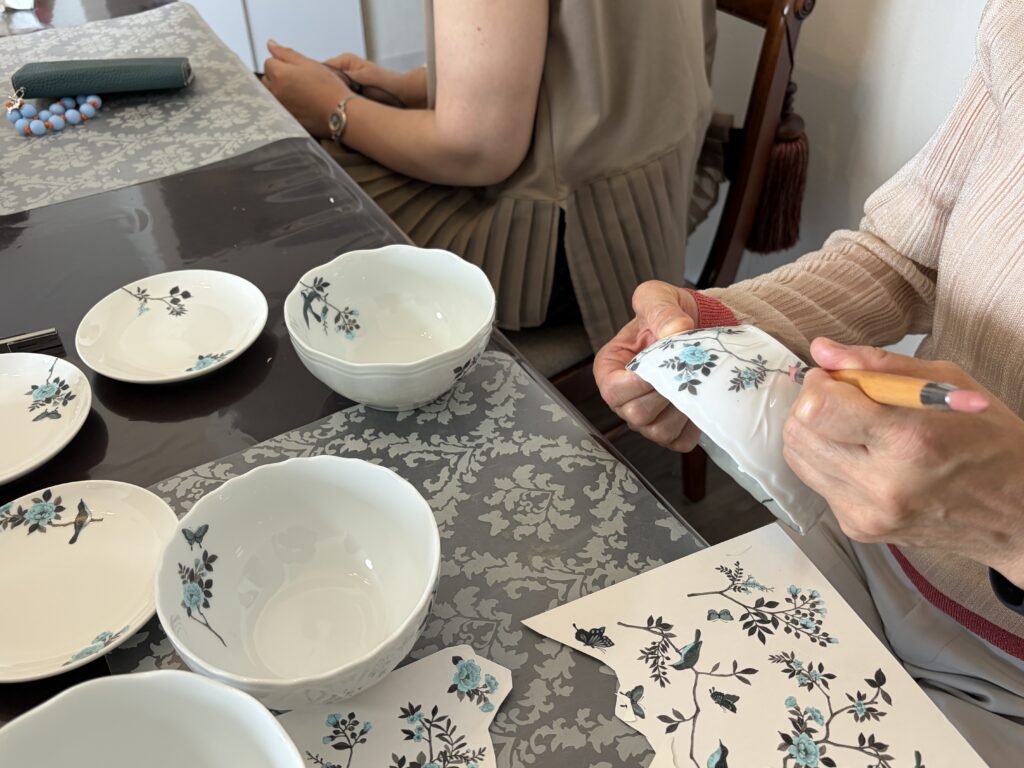 白い磁器の器に、青い花や蝶が描かれた転写紙を丁寧に貼り付けているポーセラーツ制作の風景。