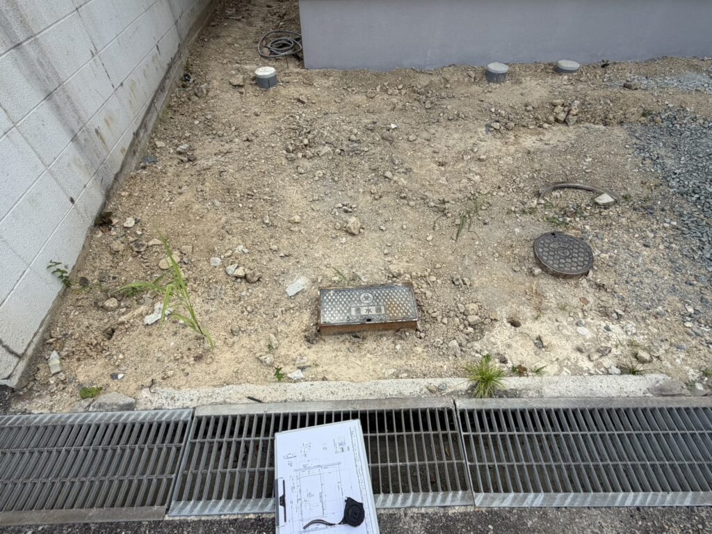 建築予定地の敷地内にて、図面やメジャーを添えて地面の状況や給水管などのインフラ位置を確認している様子。