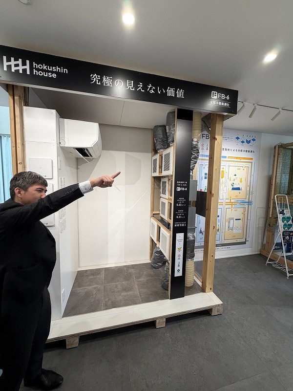 工務店の担当者がショールームで全館冷暖房システムの模型を指差し、性能や仕組みについて説明している様子。