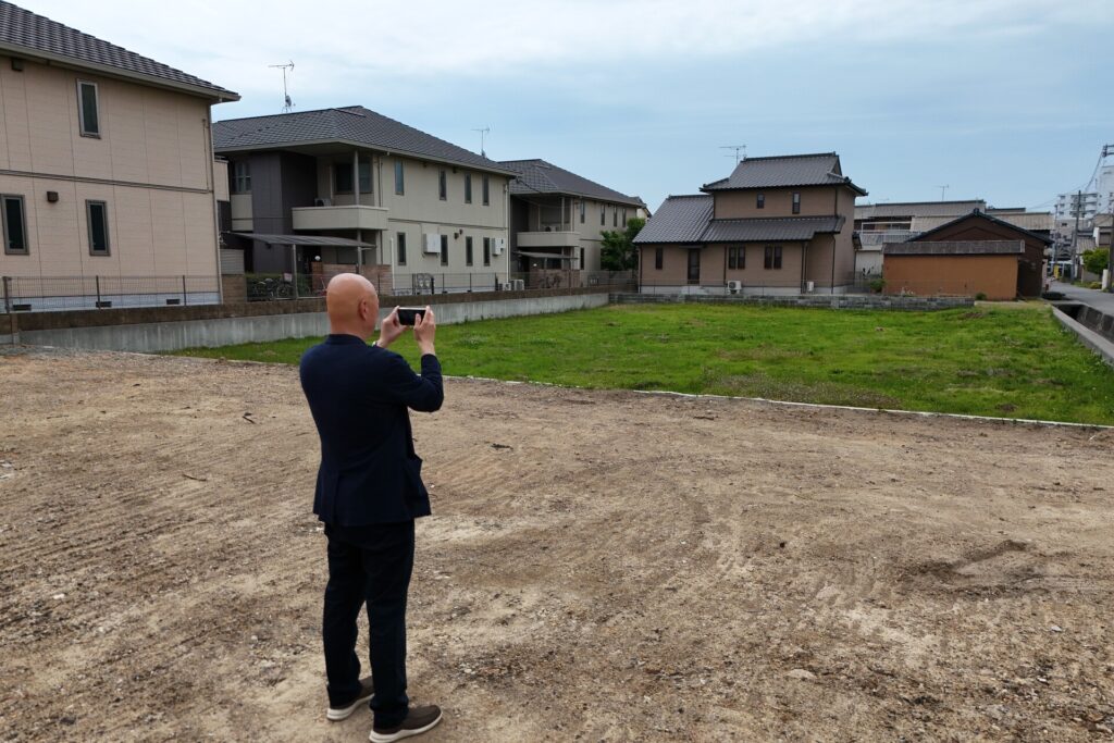 造成された更地でスマートフォンを構え、日当たりや周辺環境を確認しながら土地を撮影する男性の後ろ姿