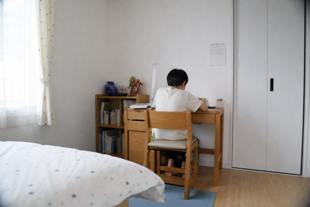 白い壁とフローリングの子供部屋で、木の温もりがある学習机に向かい勉強する男の子の後ろ姿。