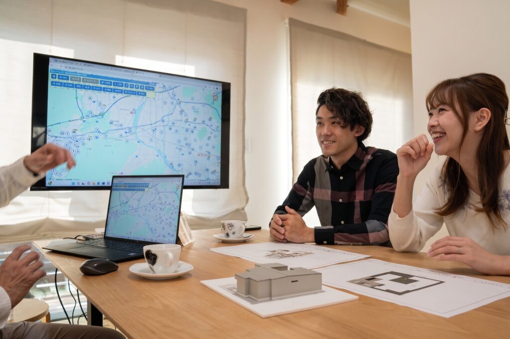 モニターに地図を表示し、テーブル上の住宅模型や間取り図面を囲んで工務店の担当者と笑顔で打ち合わせをするご夫婦。