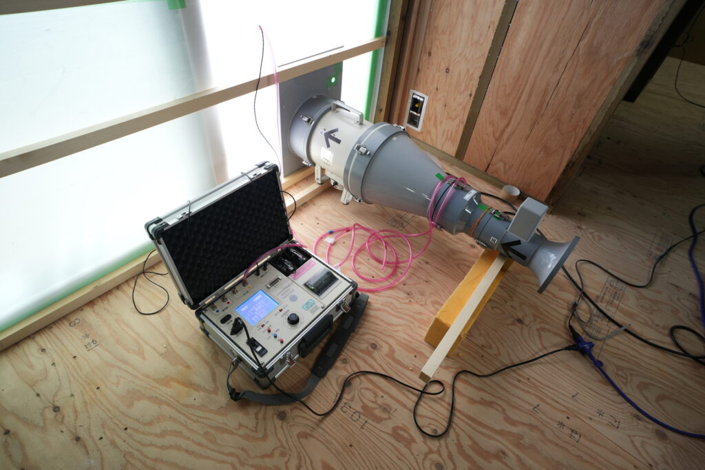 気密測定のためのブロワードア試験機が設置された室内で、建物の気密性能を確認する専門的な検査工程が写された場面。