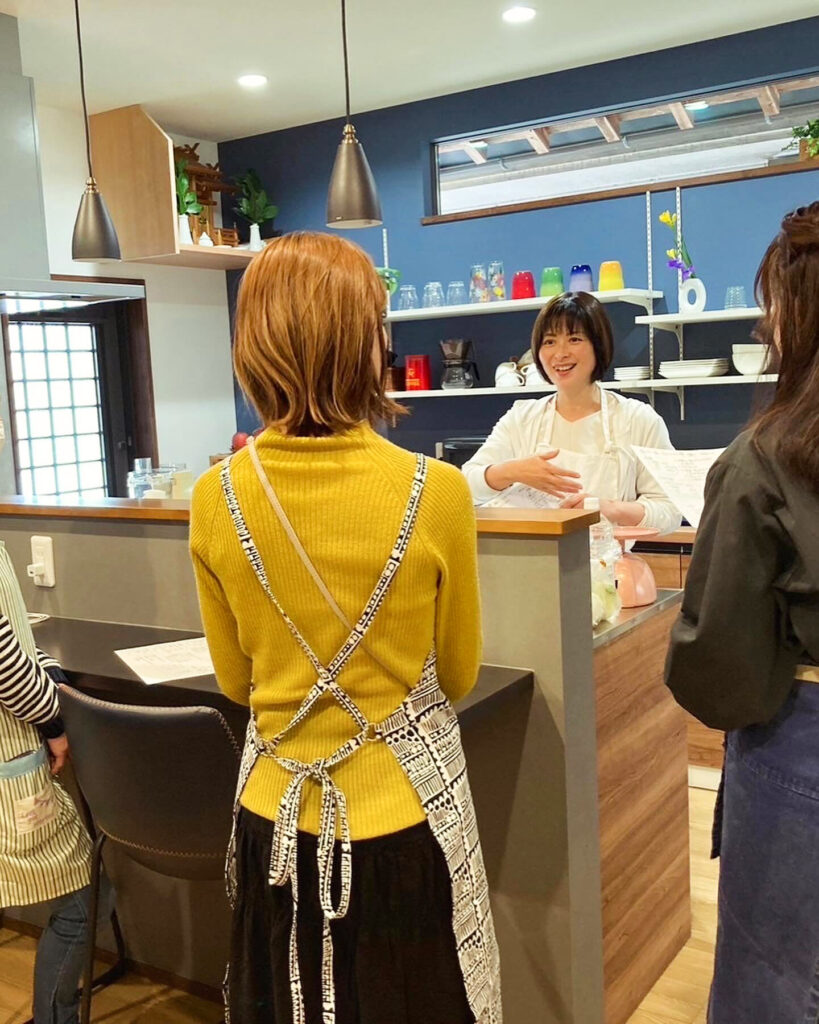 工務店の建築工房いろはが施工した、木目調のカウンターや青い壁、おしゃれなペンダントライトが特徴的なキッチンで開催された料理教室にて、エプロン姿の参加者たちが楽しそうに交流する様子。