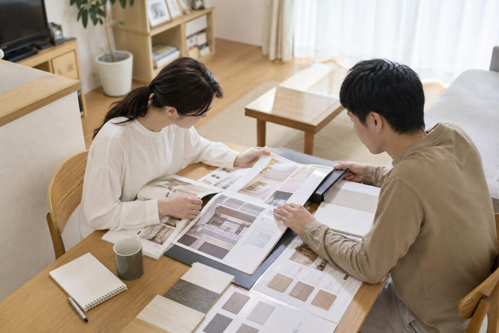 テーブルに広げた図面や資料を2人が確認しながら相談する様子が写され、住まいの計画や資金検討を行う打ち合わせ風景。