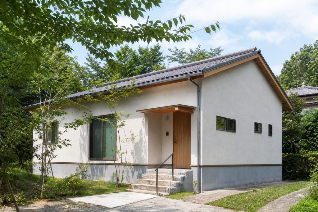 白壁に木製玄関ドアと瓦屋根を組み合わせた、植栽の緑に囲まれた平屋の外観