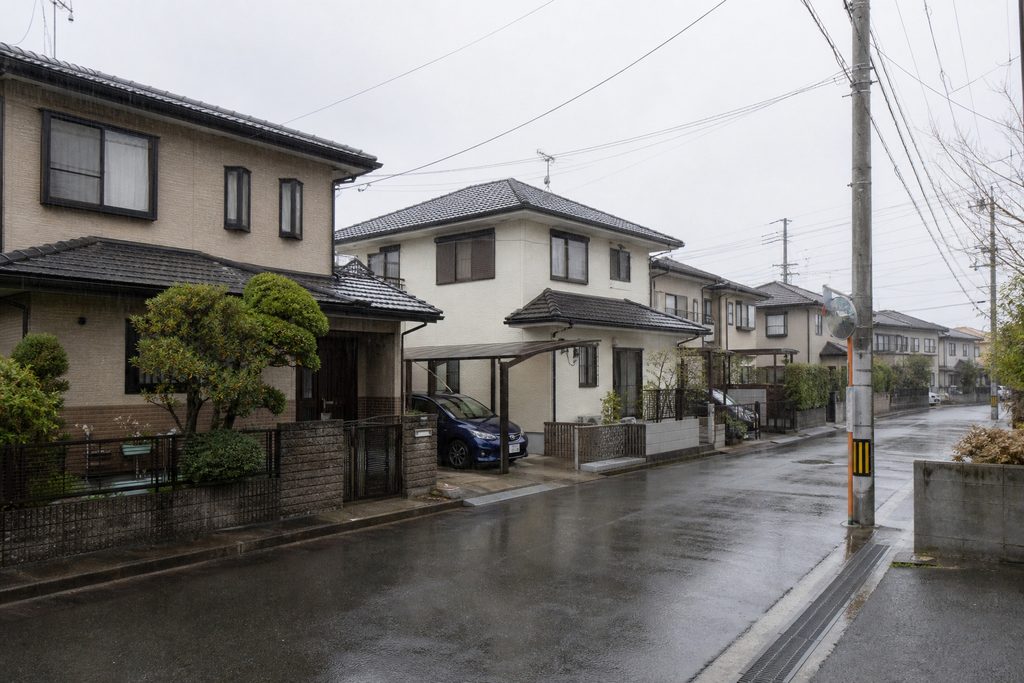 雨に濡れた閑静な住宅街の舗装道路と、左右に立ち並ぶ戸建て住宅の佇まい
