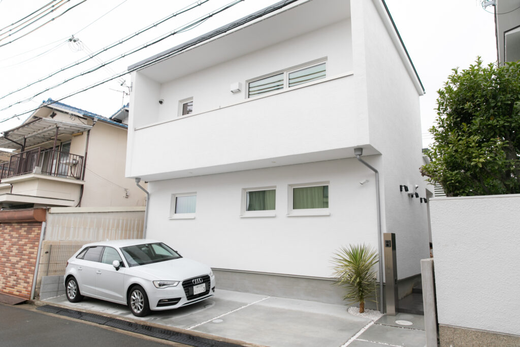 白い外壁とバルコニーが印象的なシンプルモダン住宅の外観と、コンクリート舗装された1台分の駐車スペース。