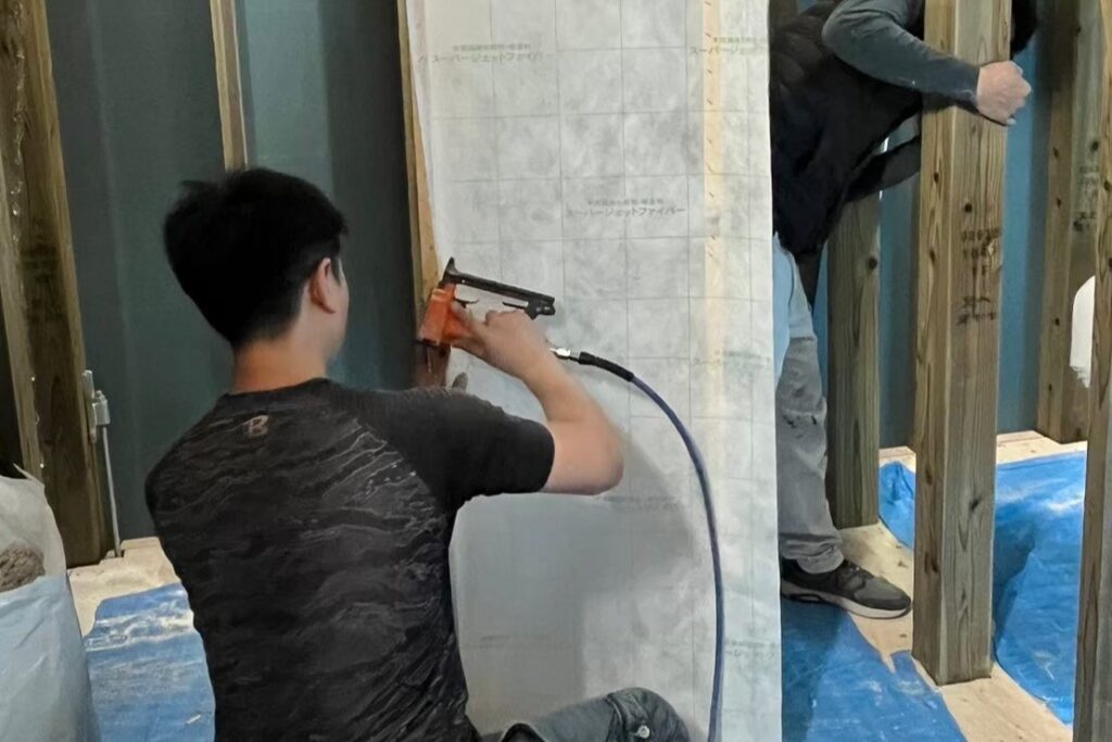木造住宅の室内で断熱材の施工や下地張りを行う作業風景で、職人が工具を使いながら建築工程を進める様子。