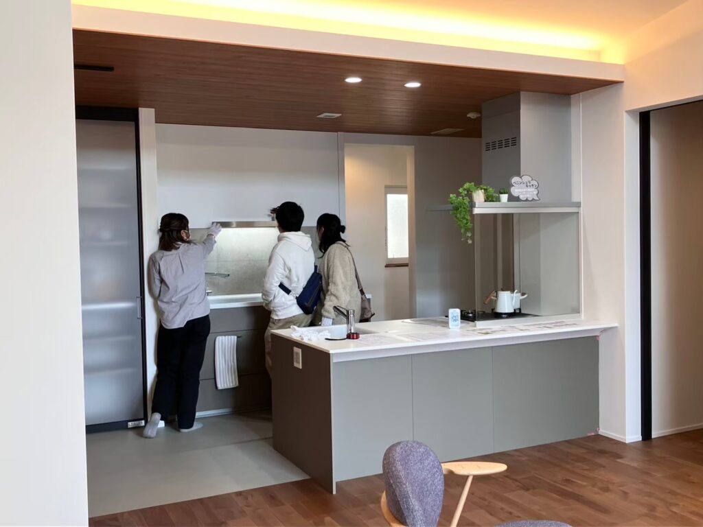明るいキッチンで3人が設備や仕上げを確認しながら話し合う様子が写され、住まいの仕様選定を行う内覧・相談の場面。