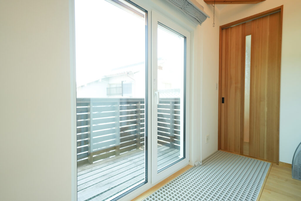 大きな掃き出し窓からウッドバルコニーへ続く明るい室内で、木製建具と床材が調和した開放的な住空間。