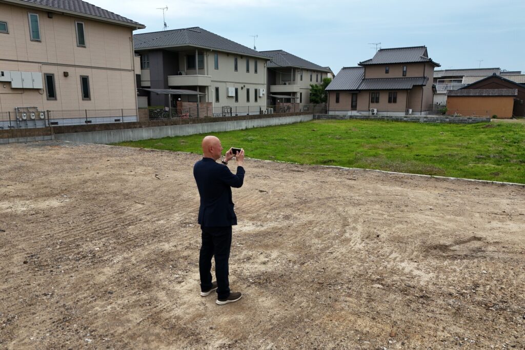 住宅街に隣接する整地された広い建築予定地で、スマートフォンを使って敷地の現況を撮影するスタッフ
