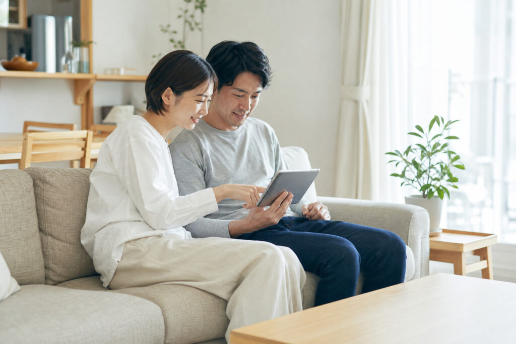 リビングのソファで仲良く並び、タブレット端末を一緒に操作しながら注文住宅やマイホームの家づくりについて情報収集や相談をする夫婦の様子。