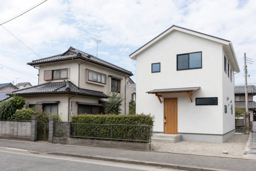 既存の住宅と隣接して建つ、木目調ドアと白い外壁が特徴的な新築一戸建ての外観を捉えた、分筆や敷地活用による注文住宅の建築例。
