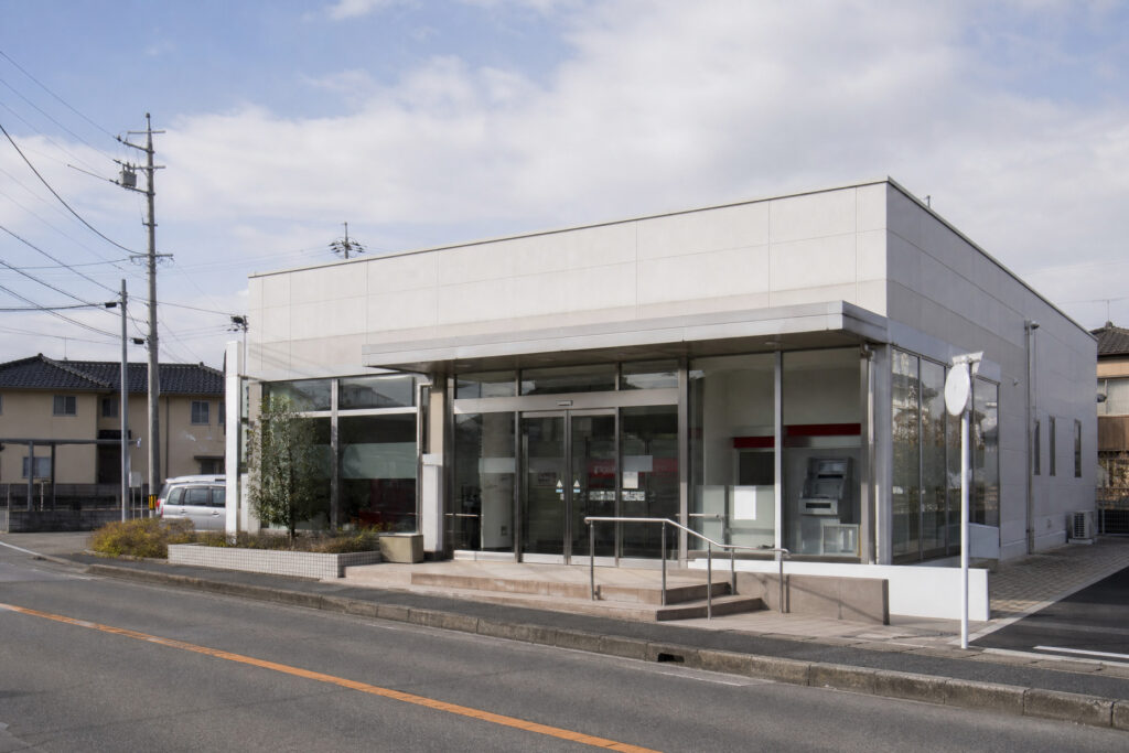 青空のもと建つ平屋風のモダンな店舗・事務所建物の外観と、ATMや店舗入り口へと続くスロープが整えられた駐車場の風景。