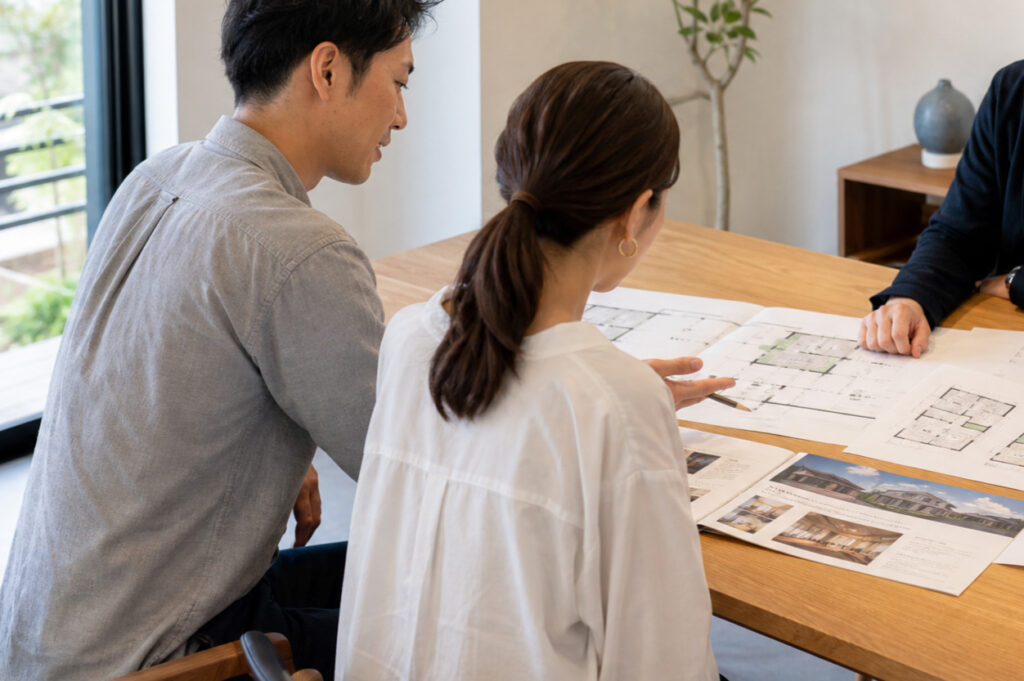机の上に広げられた住宅の間取り図面や建築カタログを見ながら家づくりや設計についての相談を行う夫婦と担当者の風景写真。
