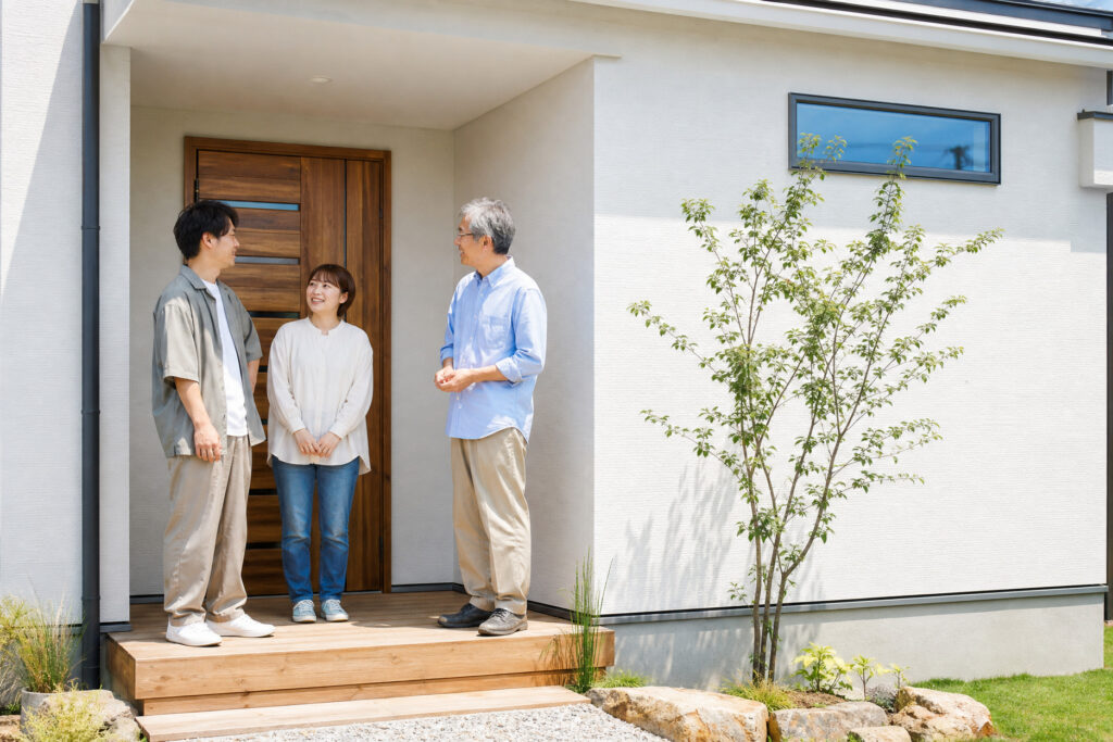 白を基調としたモダンな注文住宅の玄関前で、家族が笑顔で会話を交わしている、安心感のある家づくりや暮らしを表現した写真。